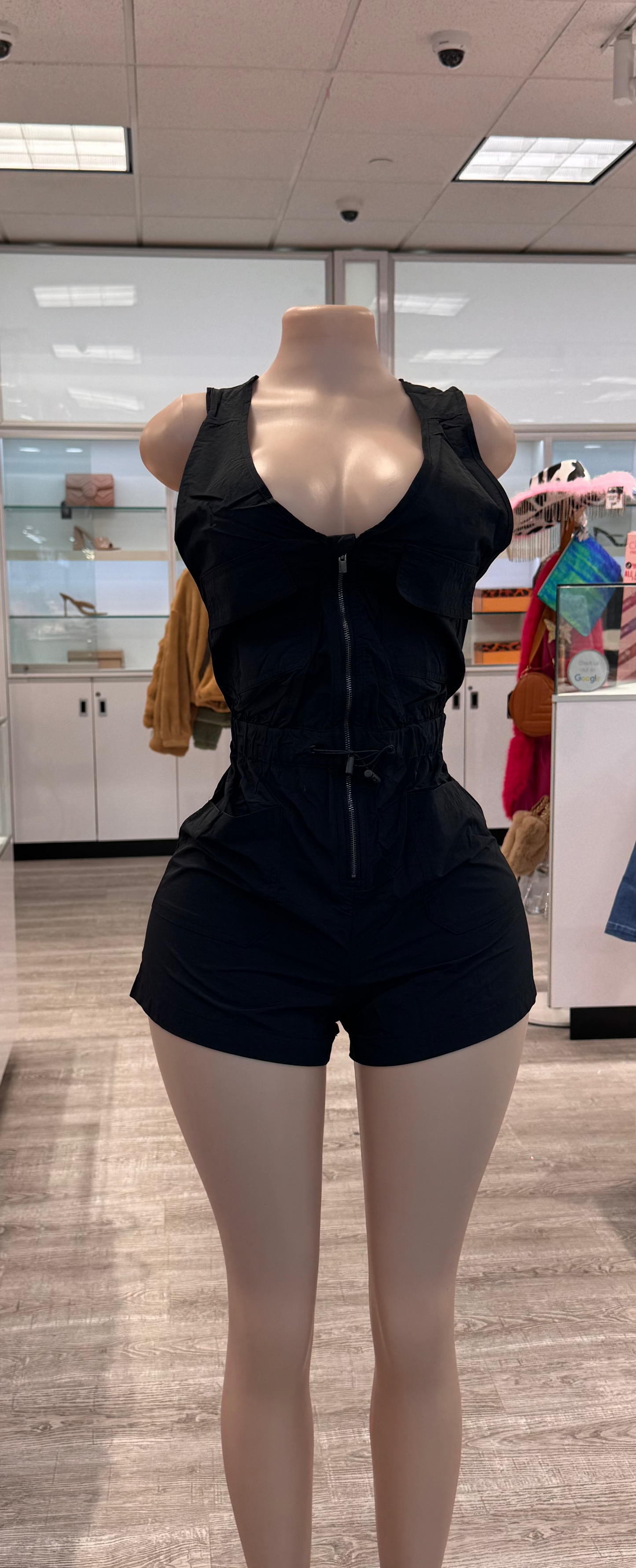 A stylish black criss-cross romper displayed on a mannequin, featuring a zip closure and adjustable drawstring waist. The romper has a fitted bodice and short legs, set against a modern retail background with various accessories visible.
