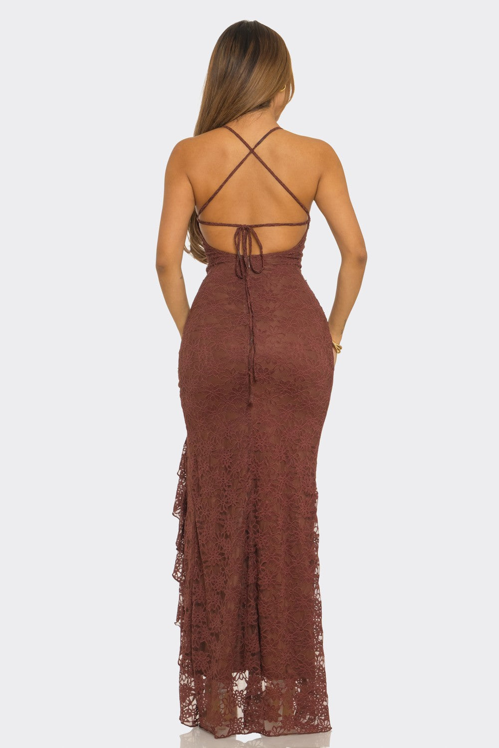 A woman viewed from the back wearing a long lace maxi dress in a muted reddish-brown color, with thin straps and a tie at the back, against a light gray studio background.