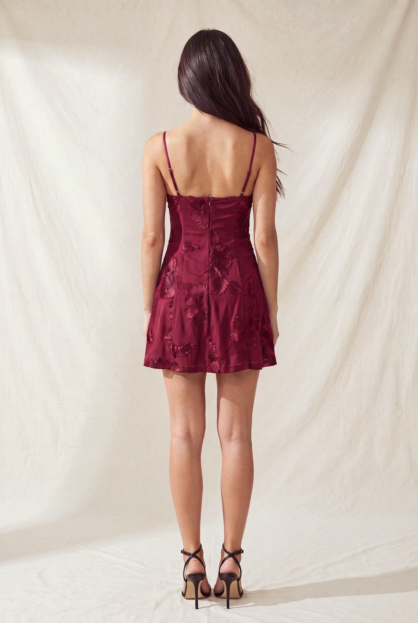 Back view of a woman in a burgundy floral dress with thin straps, standing on a light textured background.