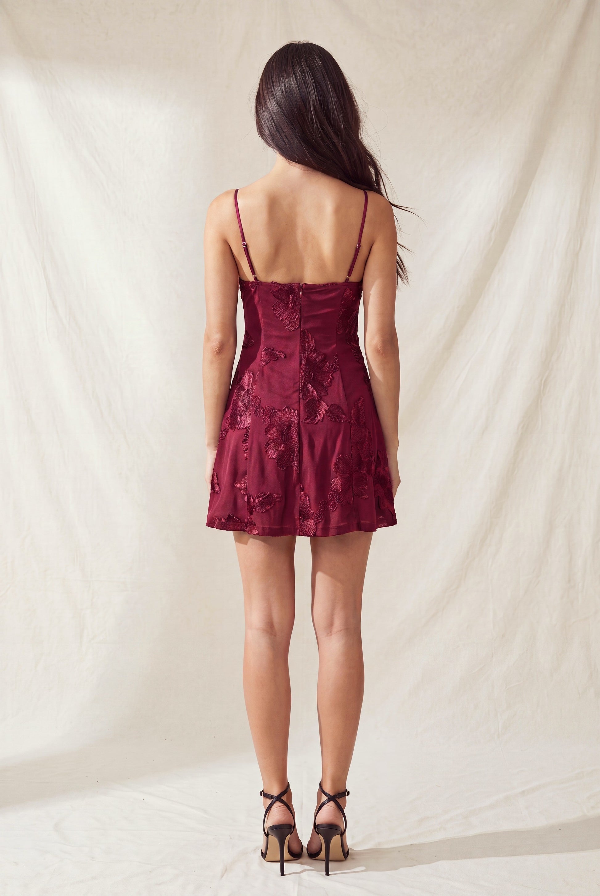 Back view of a woman in a burgundy floral dress with thin straps, standing on a light textured background.