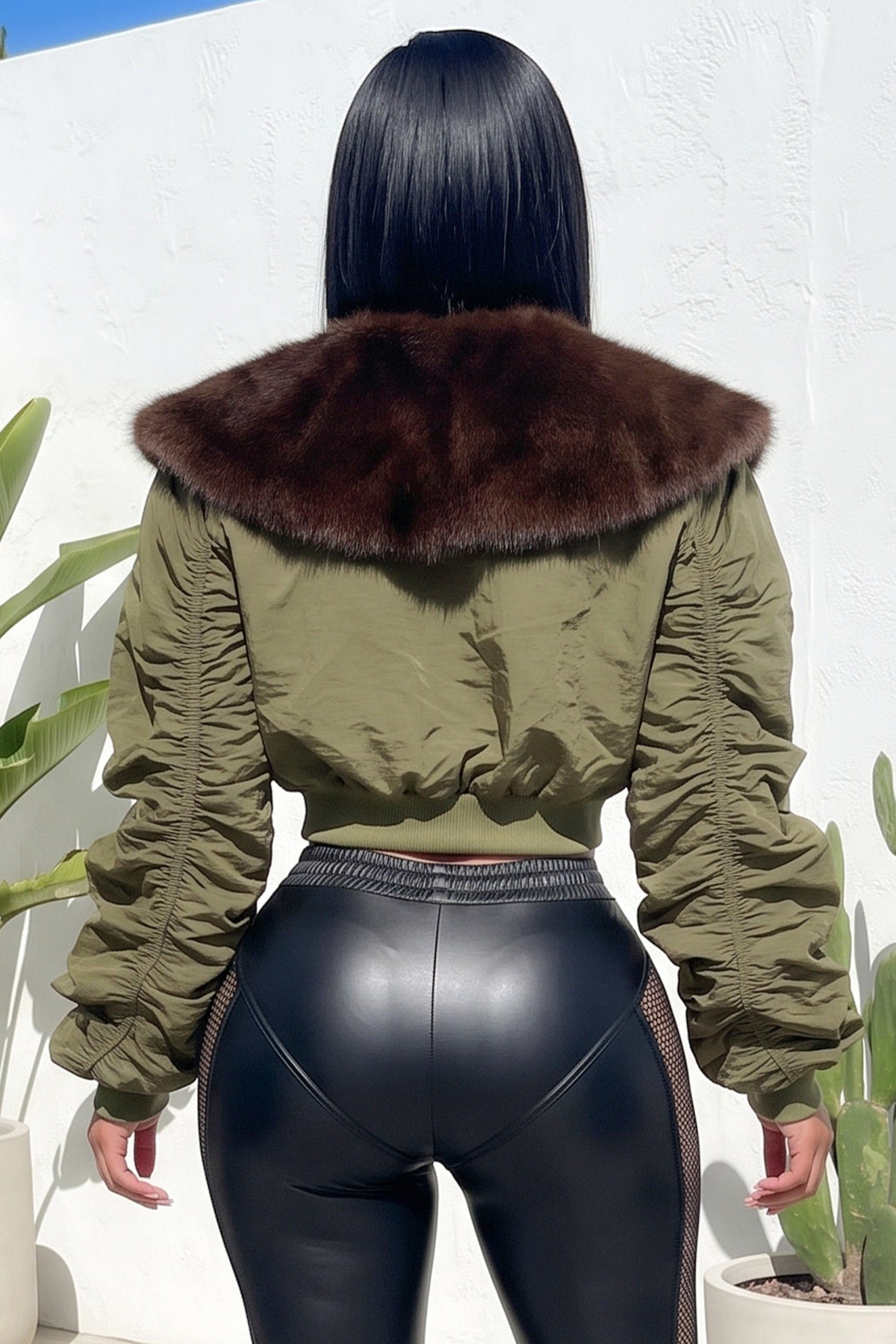 A woman shown from behind wearing an olive green cropped jacket with a large dark brown fur collar, paired with black leather-look leggings, standing against a white wall.