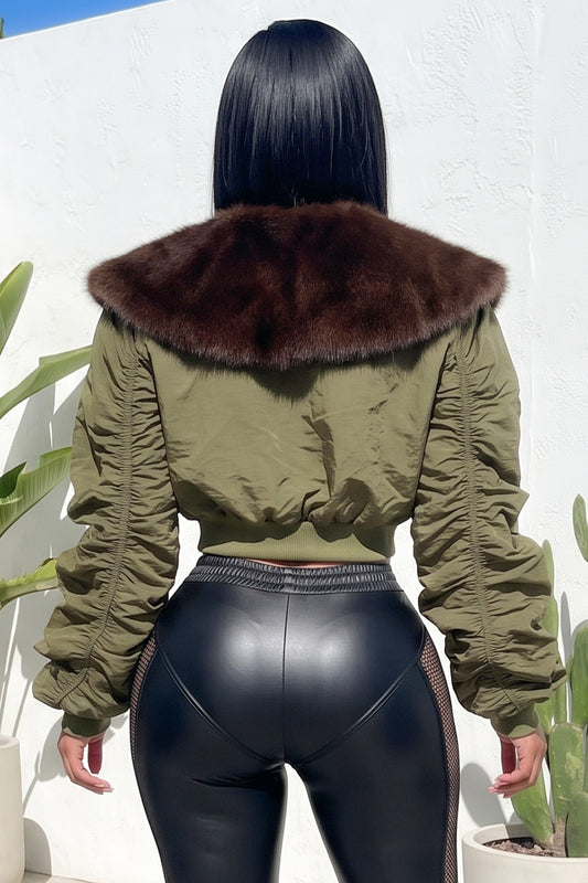 A woman shown from behind wearing an olive green cropped jacket with a large dark brown fur collar, paired with black leather-look leggings, standing against a white wall.