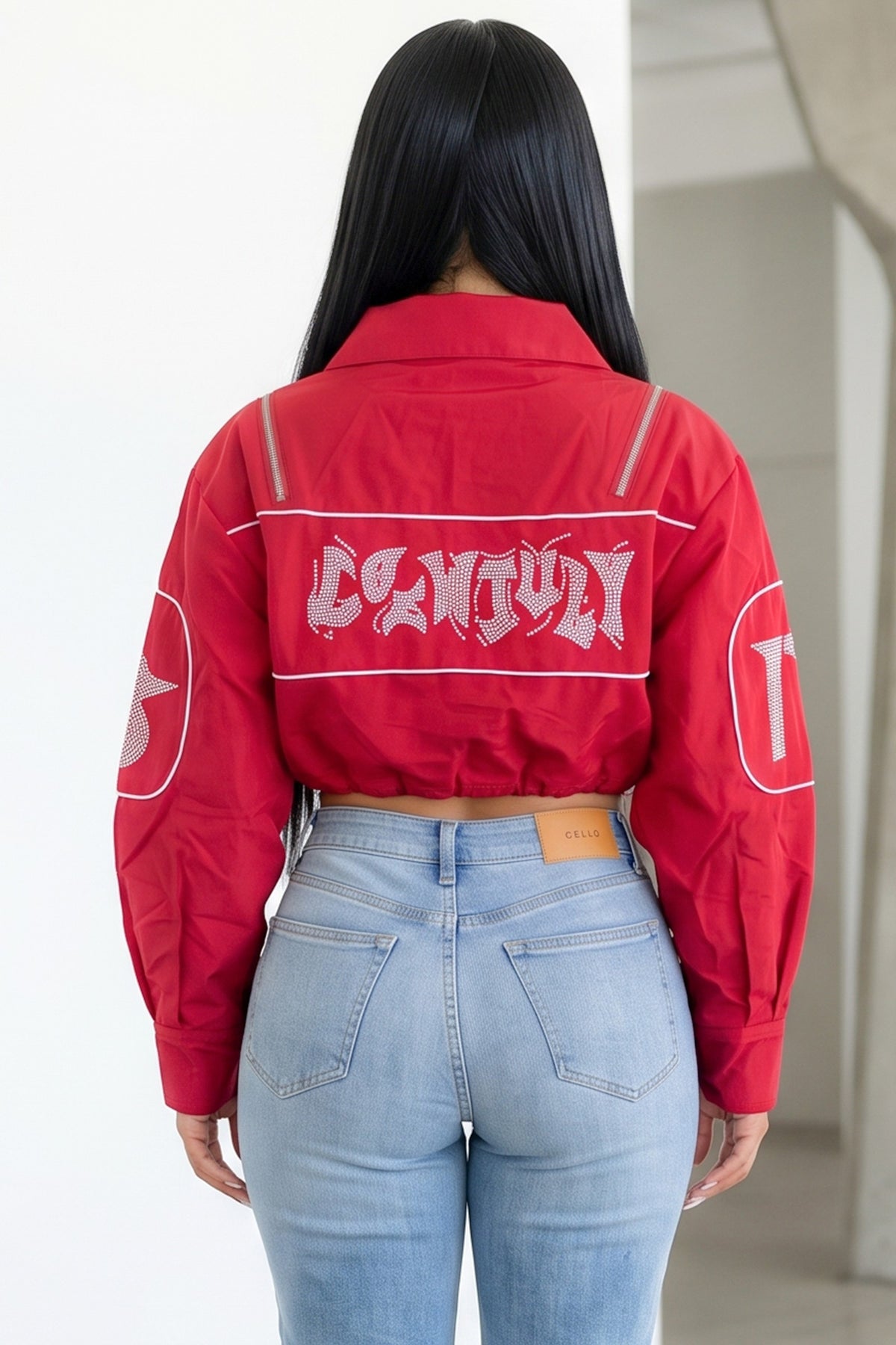 A woman viewed from behind wearing a bright red cropped jacket with a large white graphic text across the back and light-wash blue jeans.