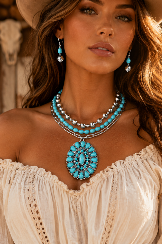 A close-up of a woman wearing a bohemian-style turquoise and silver jewelry set, featuring a large oval pendant necklace, matching drop earrings, and a multi-strand necklace, paired with a cream off-the-shoulder top.