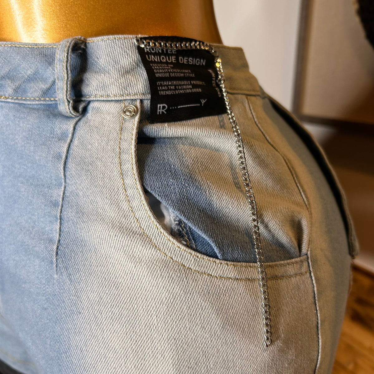 Close-up of a pair of light-wash denim jeans on a gold mannequin torso, showing the back pocket with a decorative silver chain hanging from the waistband and a black brand label.