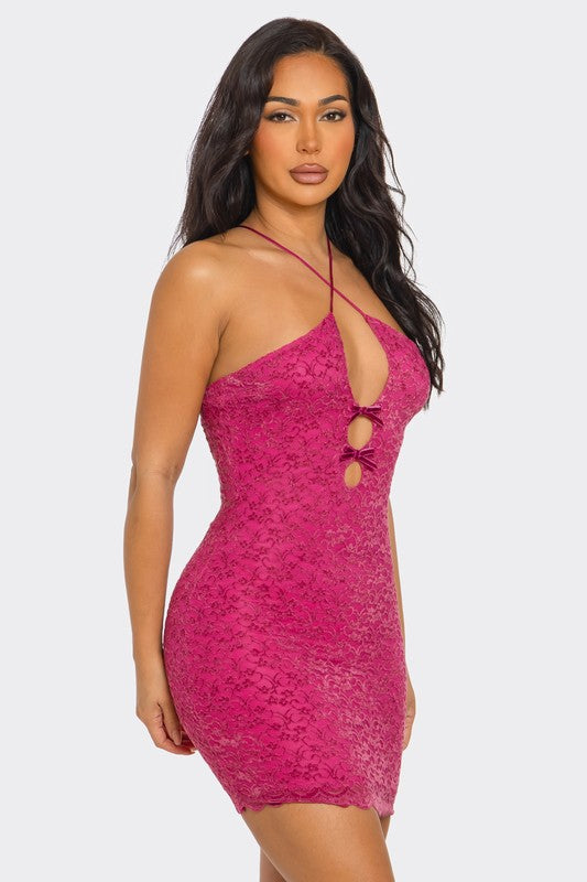 A woman in a vibrant fuchsia mini dress with a cutout bow design, featuring a plunging neckline and strappy details. Her long dark hair cascades over her shoulders as she poses confidently.