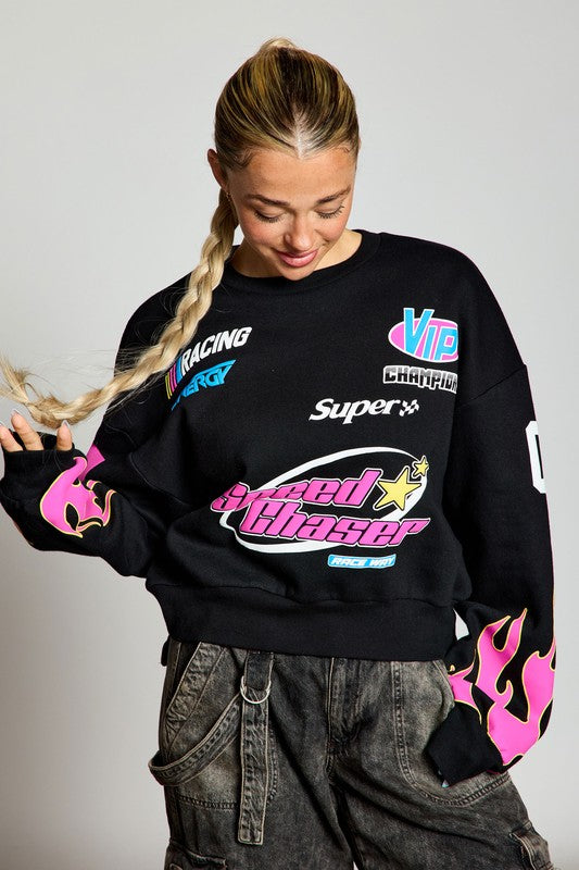 A model wearing a black sweatshirt with pink flame designs on the sleeves. The sweatshirt features colorful racing graphics including 'Speed Chaser' and brand logos, paired with casual gray pants.