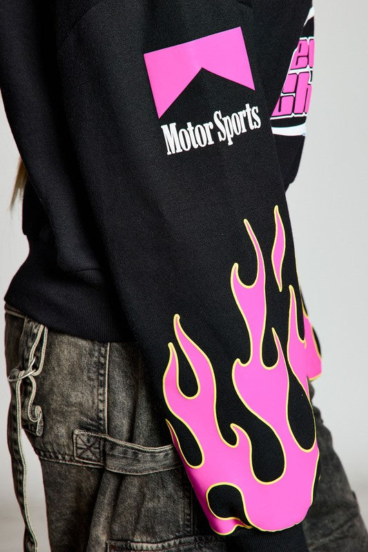 Detail view of the sleeve of a black sweatshirt, showing pink flames and the text 'Motor Sports'. The image captures the trendy design elements of the garment.