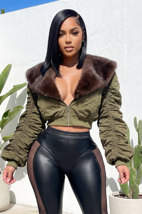 A woman modeling an olive green cropped bomber jacket with a large brown faux fur collar, paired with black mesh-paneled leggings, standing against a white stucco wall with potted cacti.