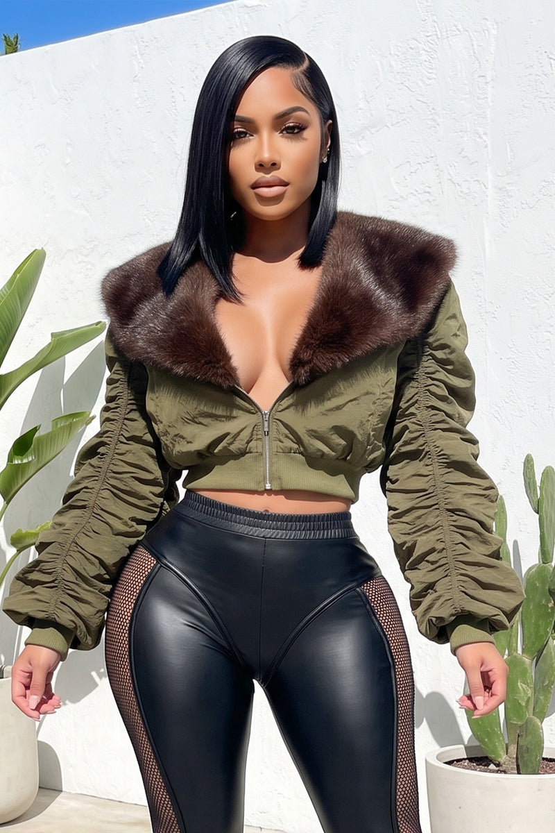 A woman modeling an olive green cropped bomber jacket with a large brown faux fur collar, paired with black mesh-paneled leggings, standing against a white stucco wall with potted cacti.