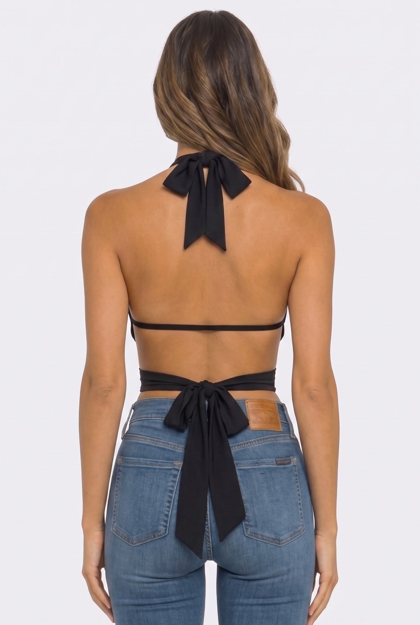 Rear view of a woman wearing a black halter-style back top with a large bow at the back of the neck and waist, paired with blue jeans, against a light gray studio background.