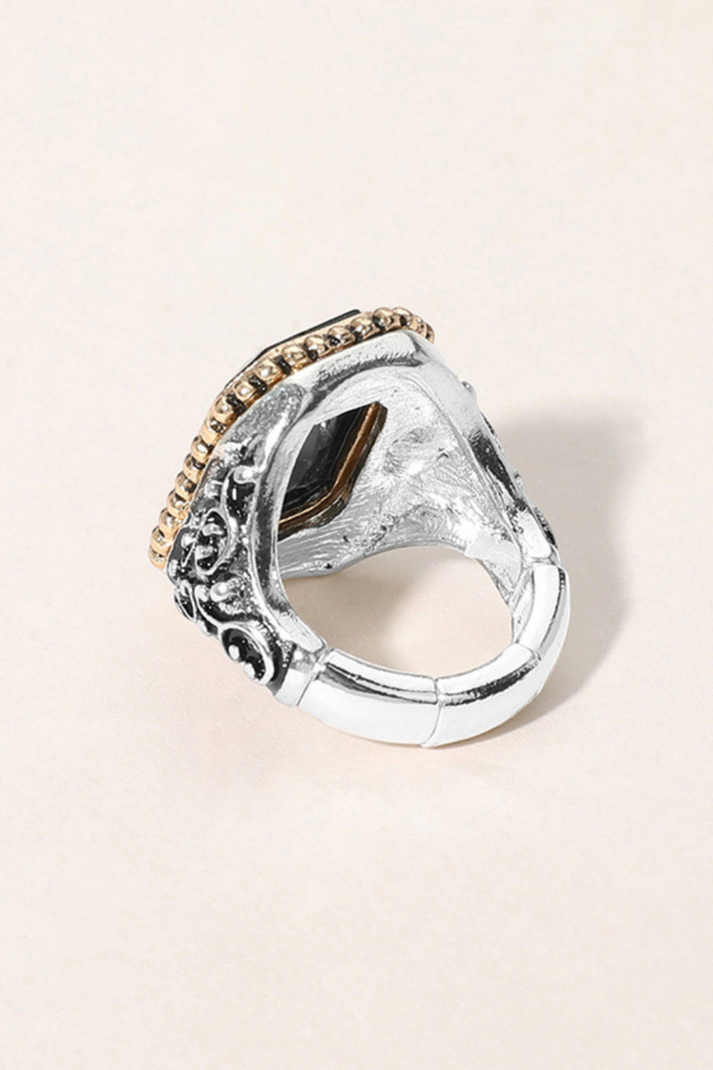 A silver-toned statement ring with an ornate carved band, a textured central face, and a row of small gold stones along the top edge, photographed on a light beige background.