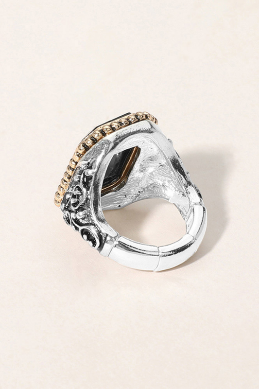 A silver-toned statement ring with an ornate carved band, a textured central face, and a row of small gold stones along the top edge, photographed on a light beige background.